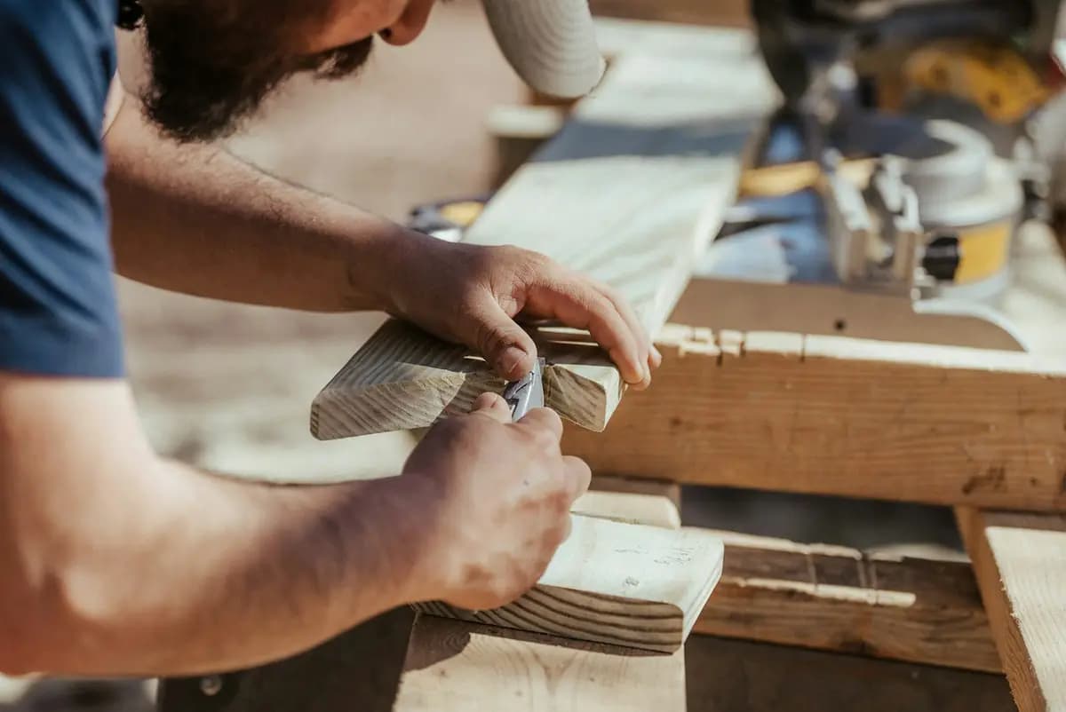 Carpenter marking a pressure-treated deck board for a cut, with a miter saw in the background