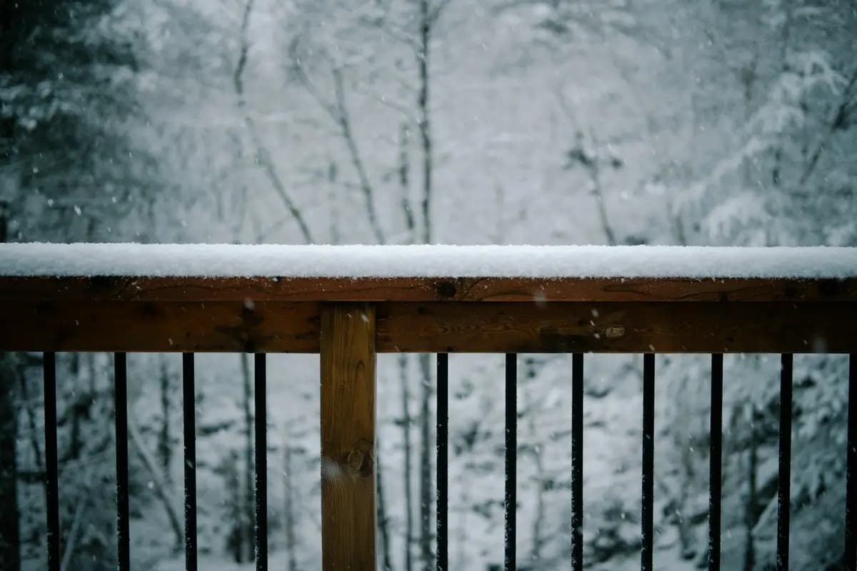 Snow resting on a wooden deck railing with black metal balusters, falling snow in the background