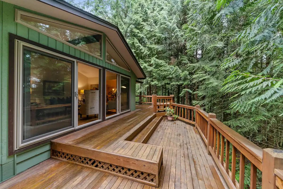 Compact wood deck with a wood railing outside sliding glass doors, surrounded by evergreens